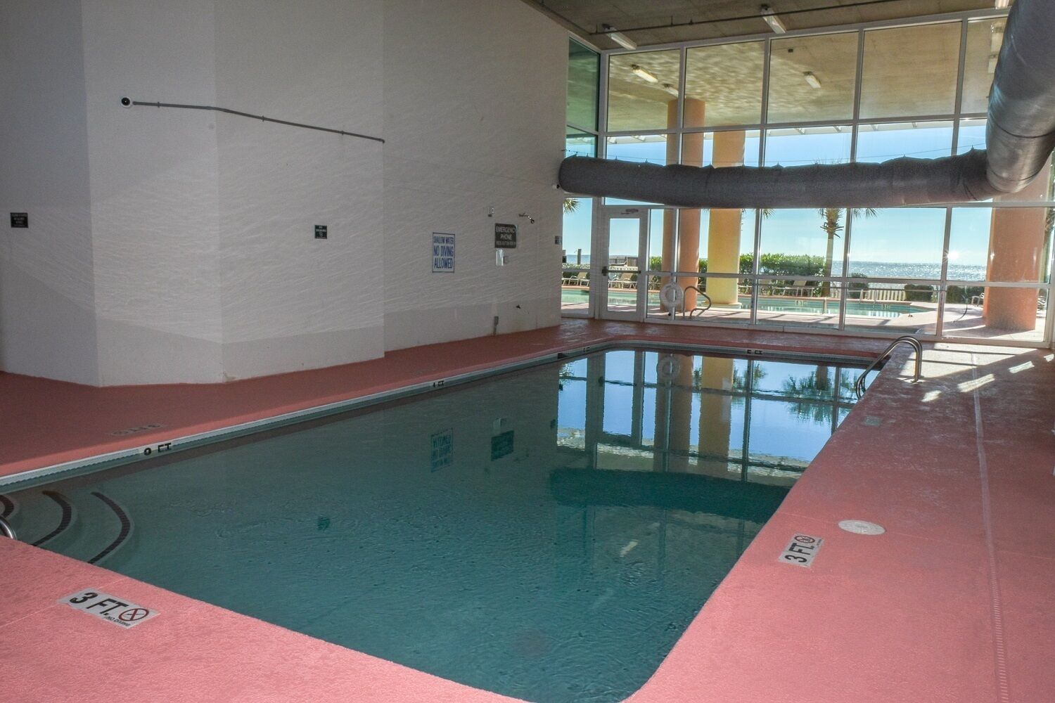 An indoor swimming pool with clear glass walls, a red floor, and a view of the outdoors.