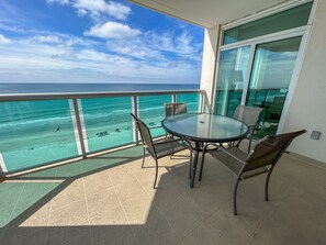 Un balcón con mesa y sillas de vidrio, con vista a una playa y al océano.