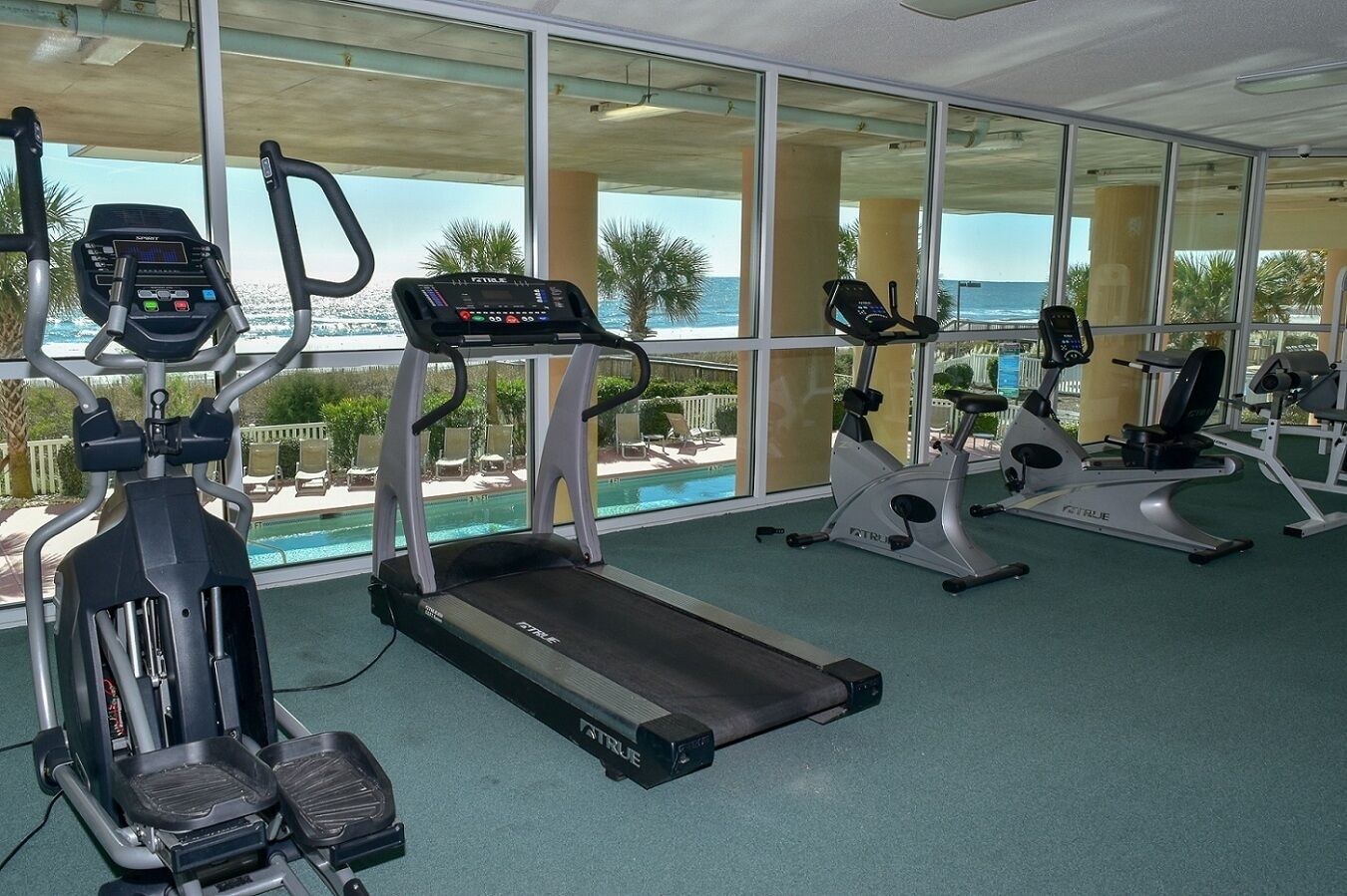 Ett gym med löpband, crosstrainers och en motionscykel, med utsikt över en pool och havet.