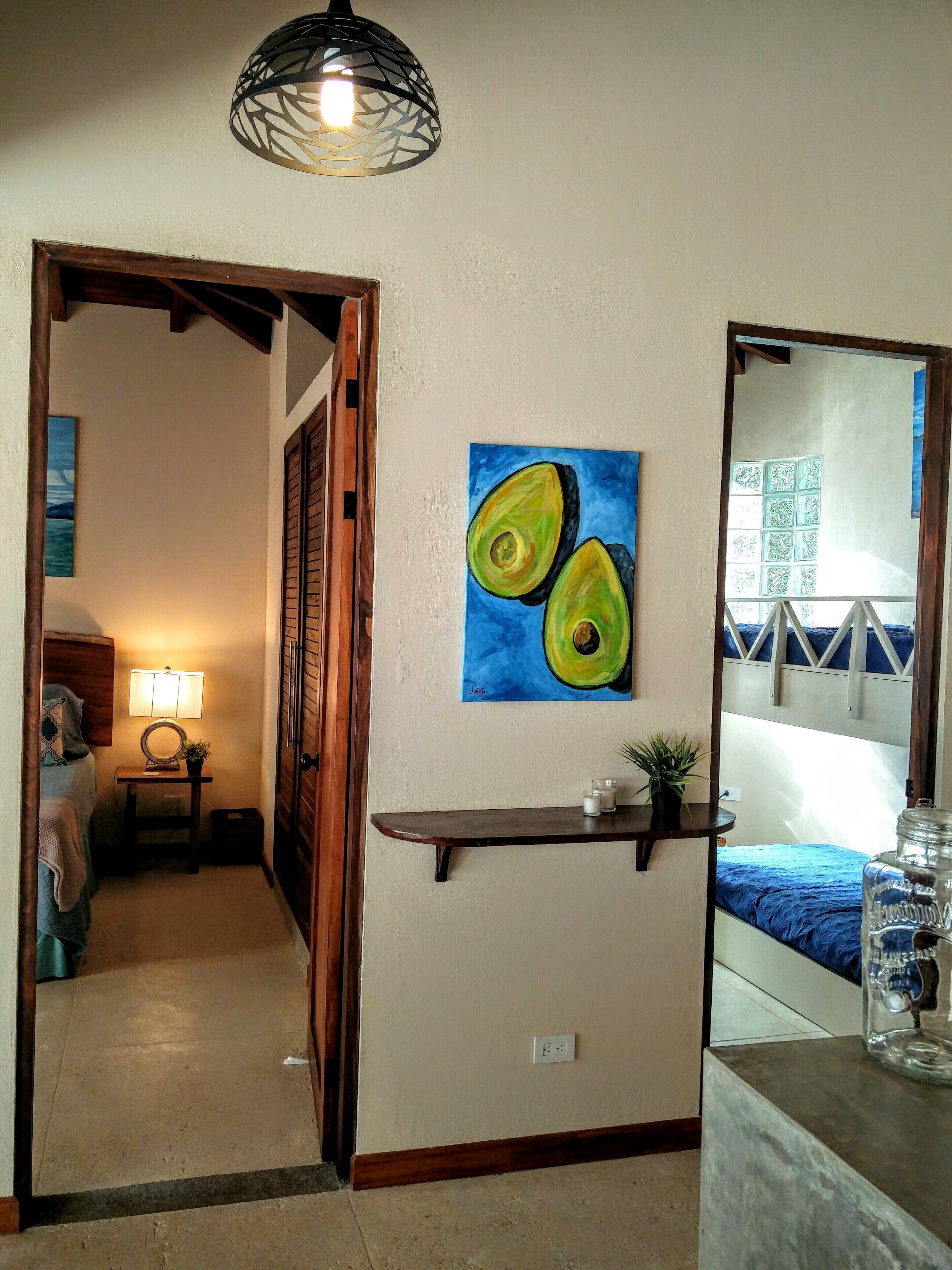 Ein Zimmer mit einem Bett, einem Nachttisch mit Lampe, einem Avocado-Gemälde, einem Spiegel und Blick ins Freie.