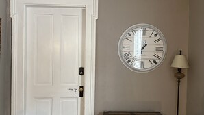 A room with a white door, a round wall clock, a wooden chest, and a floor lamp.