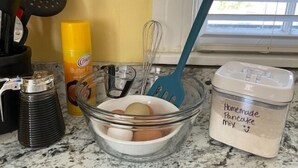 A kitchen counter with a glass bowl containing eggs, a yellow bottle, a blue spatula, and a container labeled "Homemade Pancake mix".