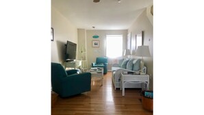 A spacious living room with a wooden floor, a teal armchair, a white sofa, and a television. - Bungalow by the Bay (Villas)