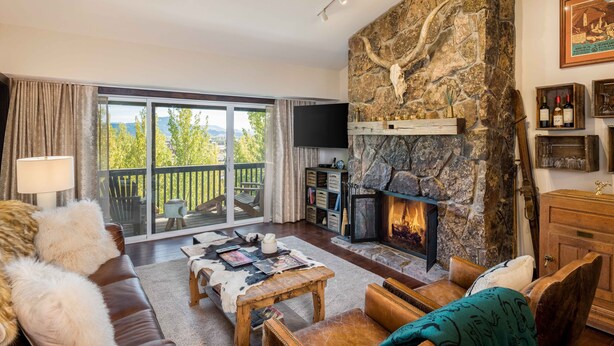 A cozy living room with a stone fireplace, a large window with a view, and a flat-screen TV. - Teton Village 3.5BR/2.5BA  - Walking Distance to Ski Lifts! (Jackson)