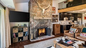 A modern living room with a stone fireplace, wooden shelves, a flat-screen TV, leather furniture, and a kitchen area. - Teton Village 3.5BR/2.5BA  - Walking Distance to Ski Lifts! (Jackson)