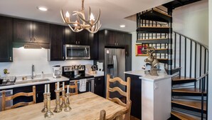 A modern kitchen with dark cabinets, stainless steel appliances, a wooden dining table, and a staircase with a metal railing. - Teton Village 3.5BR/2.5BA  - Walking Distance to Ski Lifts! (Jackson)