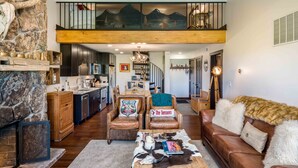A spacious living room with a stone fireplace, leather furniture, and a high ceiling with a balcony. - Teton Village 3.5BR/2.5BA  - Walking Distance to Ski Lifts! (Jackson)