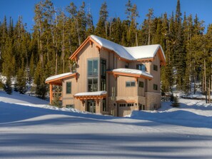 Une cabane en bois à plusieurs étages, dotée de grandes fenêtres et d’un toit recouvert de neige, entourée d’une forêt.