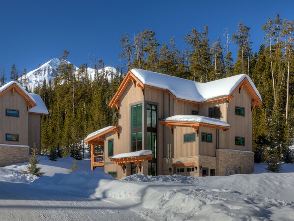Un chalet en bois à plusieurs étages, dont le toit est recouvert de neige, entouré d’arbres et avec une montagne en arrière-plan.