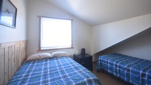 Un dormitorio con cabecera de madera, un ventanal grande, una mesita de noche y una cama litera con colcha azul a cuadros.