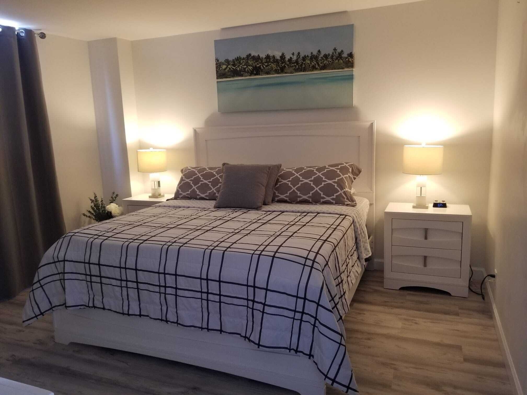 Un dormitorio con una cama, dos mesitas de noche, un aparador, un cuadro y una cortina.