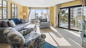 Un salón espacioso con un sofá grande, un sillón más pequeño y un comedor con mesa y sillas. Se ve un balcón con piscina a través de las puertas de cristal.