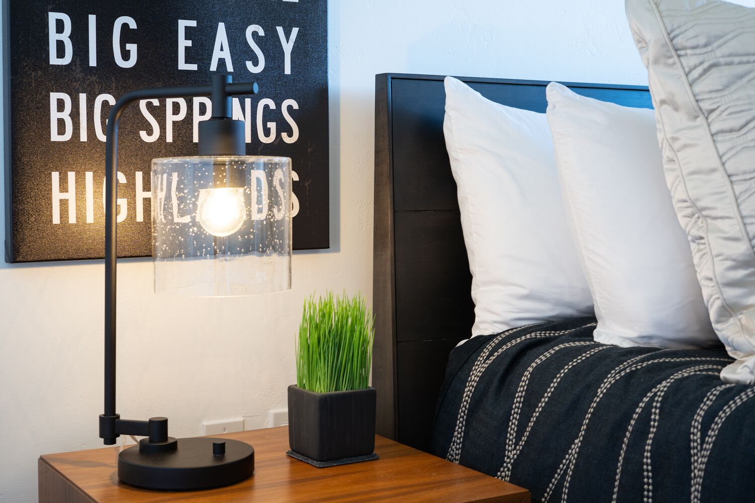 Une chambre à coucher bien rangée, avec un lit, des oreillers, une table de chevet, une lampe et un mur orné d’une citation inspirante.