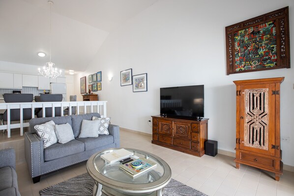 A living room with a grey sofa, a glass coffee table, a TV, a wooden cabinet, and a large painting on the wall. - *OCEANFRONT Cosy and modern style condo* (Cupecoy)