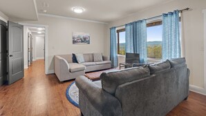 Ein modernes Wohnzimmer mit einer grauen Sofagarnitur, einem Couchtisch und einem Fernseher. Ein Fenster mit blauen Vorhängen bietet Blick ins Freie.