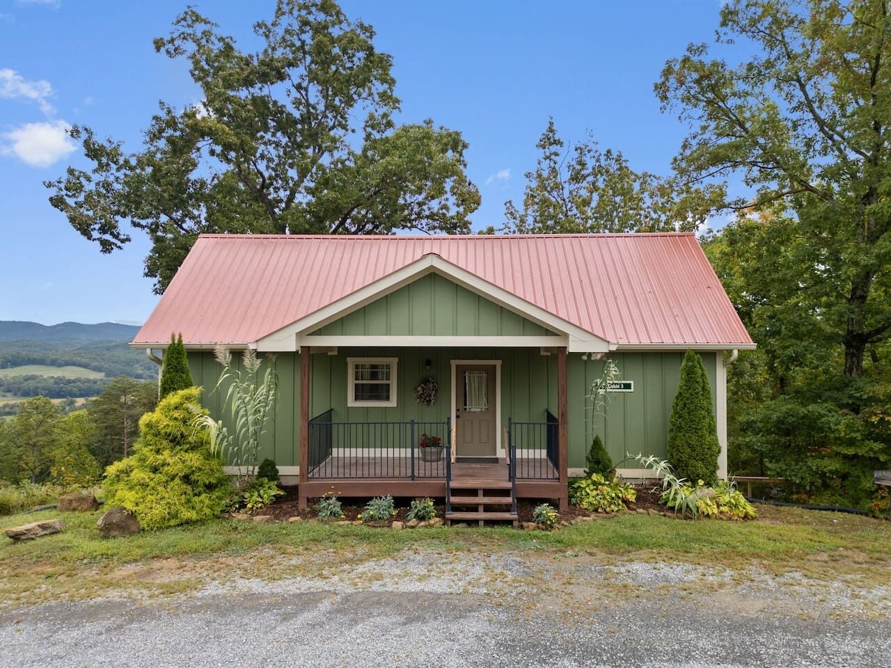 Une petite maison à un seul étage, à l’extérieur vert et au toit rouge, entourée d’une végétation luxuriante et d’arbres.
