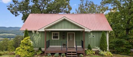 Une petite maison à un seul étage, à l’extérieur vert et au toit rouge, entourée d’une végétation luxuriante et d’arbres.