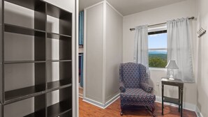 Ein Zimmer mit einer großen, schwarz-weißen Regaleinheit, einem gemusterten Sessel, einem kleinen Tisch mit Lampe und einem Vorhangfenster.