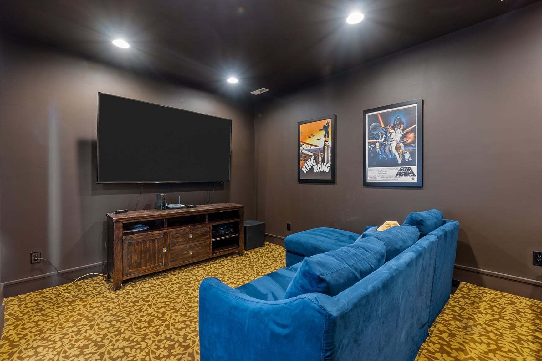 A home theater room with a large flat-screen TV, a wooden entertainment center, a blue sectional sofa, and two framed movie posters on the wall.