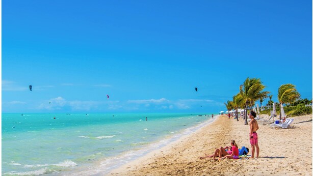 Una playa con agua turquesa cristalina, gente tomando el sol y algunos cometas volando en el cielo.