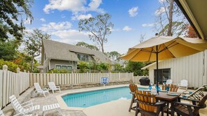 A backyard with a swimming pool, white deck chairs, a wooden table with chairs, and a covered patio area.