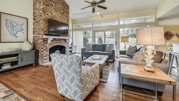 A spacious living room with a brick fireplace, a ceiling fan, and a large window offering a view of the outdoors.