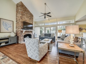 A spacious living room with a brick fireplace, a ceiling fan, and a large window offering a view of the outdoors.