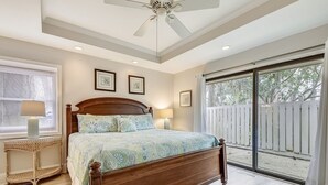 A bedroom with a wooden bed, two bedside lamps, a ceiling fan, and a large window with a view of a white fence and trees.