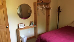A bedroom with a wooden bed frame, a round mirror, a shelf with books and a framed picture, a hanging coat rack, and a bedside table with a trash bin. - 5 BR/2.5 BA Hunting & Fishing Lodge (Watertown)