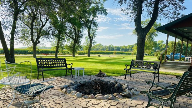 An outdoor patio with stone flooring, metal chairs, and benches, surrounded by trees and a grassy field. - 5 BR/2.5 BA Hunting & Fishing Lodge (Watertown)
