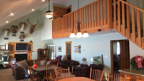 A spacious living area with a dining table, chairs, a fireplace, a television, and a loft with a wooden railing. - 5 BR/2.5 BA Hunting & Fishing Lodge (Watertown)