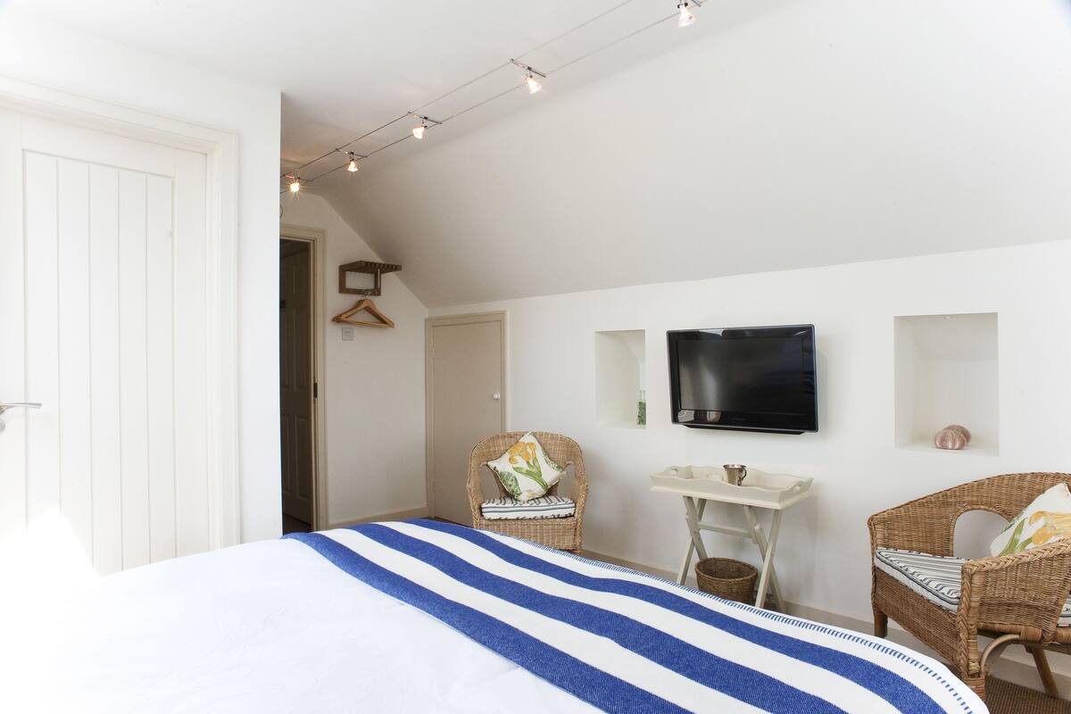 A bedroom with a bed, two wicker chairs, a small table, a TV, and a door.