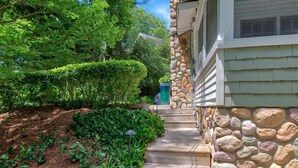 A stone-clad house with a wooden pathway leading to a front door, surrounded by lush greenery and a well-maintained garden.