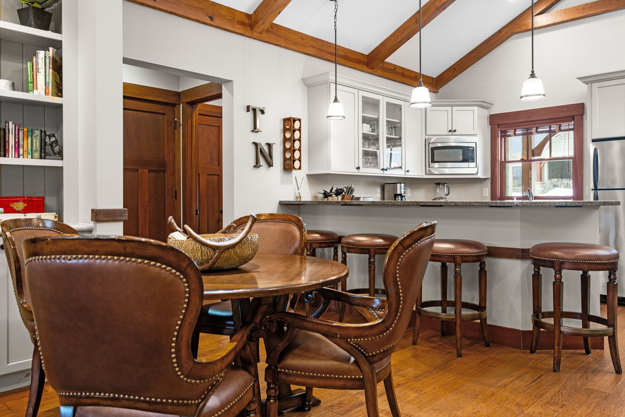 A dining area with a wooden table and chairs, a kitchen with white cabinets and a microwave, and a wooden ceiling with exposed beams.