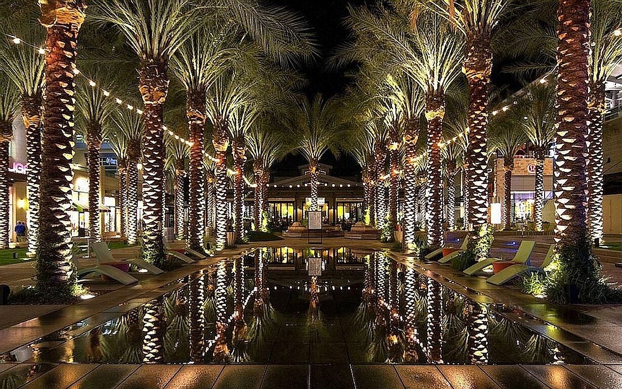 A nighttime view of a hotel corridor lined with palm trees and illuminated by string lights, leading to a central building with a reflective pool.