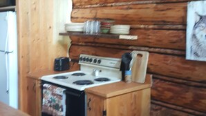 A wooden cabin kitchen with a stove, wooden cabinets, and a shelf with dishes. - Rustic cabin hidden among the pines. (Condon)
