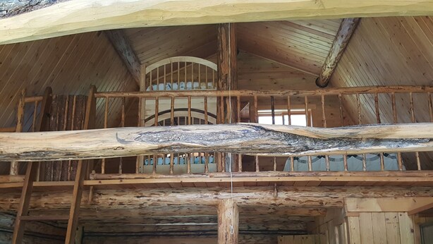 A wooden interior with a large, curved beam spanning the ceiling, supported by wooden beams and a metal railing. - Rustic cabin hidden among the pines. (Condon)