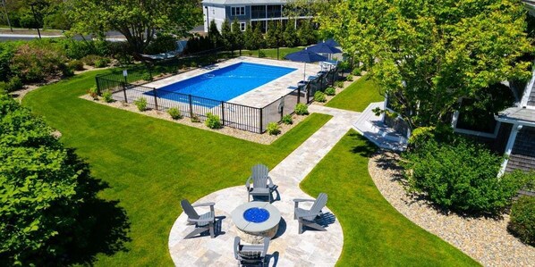 A rectangular swimming pool surrounded by a black metal fence, with a stone fire pit and four Adirondack chairs in the foreground. - A Wave From It All: 227-O Overlooks Nauset Beach/Atlantic, Heated Pool, Hot Tub & Fire-pit (Orleans)