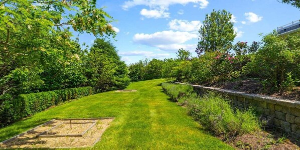 A well-maintained garden with a stone wall, neatly trimmed hedges, and a clear pathway. - A Wave From It All: 227-O Overlooks Nauset Beach/Atlantic, Heated Pool, Hot Tub & Fire-pit (Orleans)