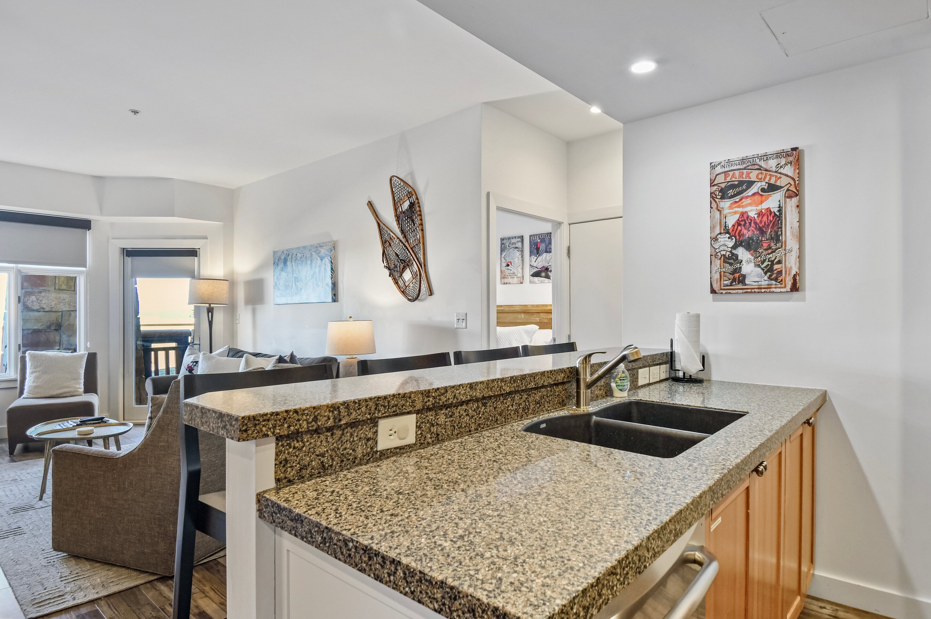 A modern kitchen with granite countertops, a sink, and wooden cabinets. There is a dining area with a table and chairs, and a living room with a sofa and a lamp.