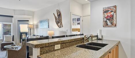 A modern kitchen with granite countertops, a sink, and wooden cabinets. There is a dining area with a table and chairs, and a living room with a sofa and a lamp.