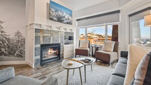 A modern living room with a fireplace, comfortable seating, and a view of the mountains through large windows. - Perfect summer! Balcony with views of the concerts, outdoor swimming pool, golf hiking and more (Park City)