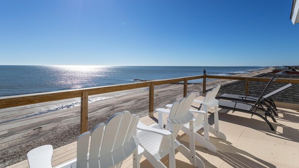 Une terrasse au bord de la plage, avec des chaises blanches et des transats, d’où l’on a une vue sur l’océan.
