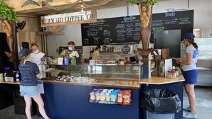 A coffee shop counter with menu boards, a trash bin, and a customer interacting with a staff member. - Newly Renovated Near Amazing Sunset Beach (Falmouth)