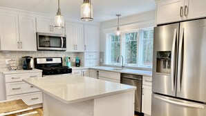 A modern kitchen with white cabinetry, stainless steel appliances, a large island with bar stools, and a window providing natural light. - Newly Renovated Near Amazing Sunset Beach (Falmouth)
