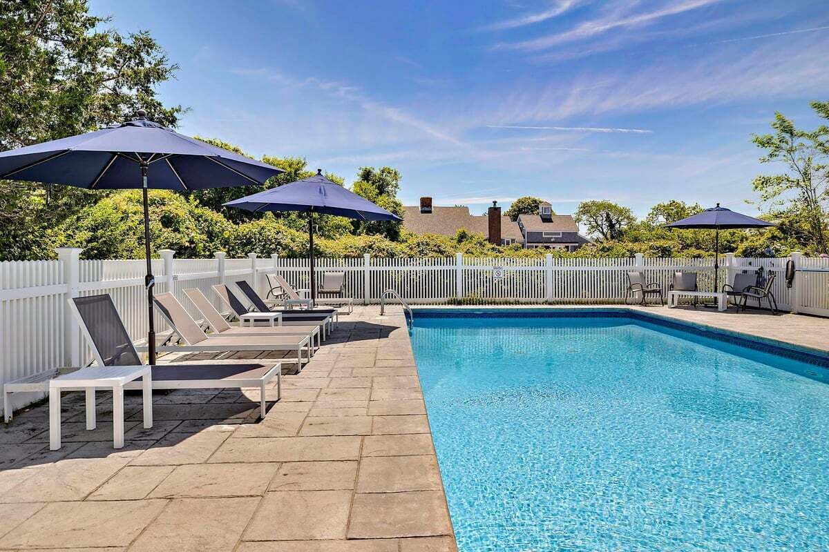 Une piscine entourée d’une clôture blanche et d’arbres, avec des chaises longues et des parasols.