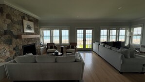 A spacious living room with a stone fireplace, a large gray sofa, and a view of the beach through large windows. - Brand new Five bedroom beachfront home on Moody Beach...Amazing location! (Wells)