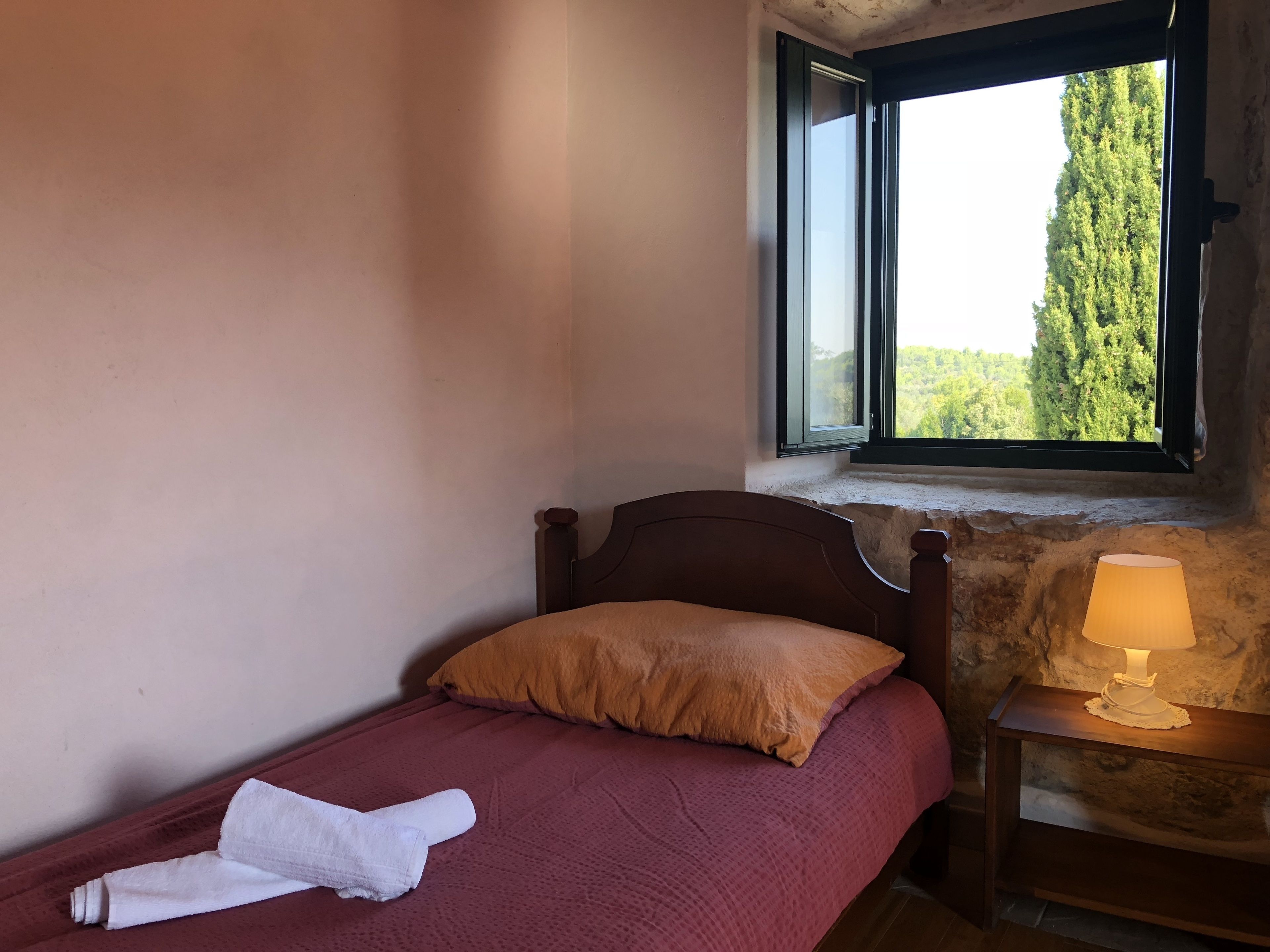Una cama individual con colcha roja, almohada marrón y toalla blanca. A la derecha de la cama hay una mesita de noche con una lámpara. Se ve una ventana con vistas a árboles por encima de la cama.