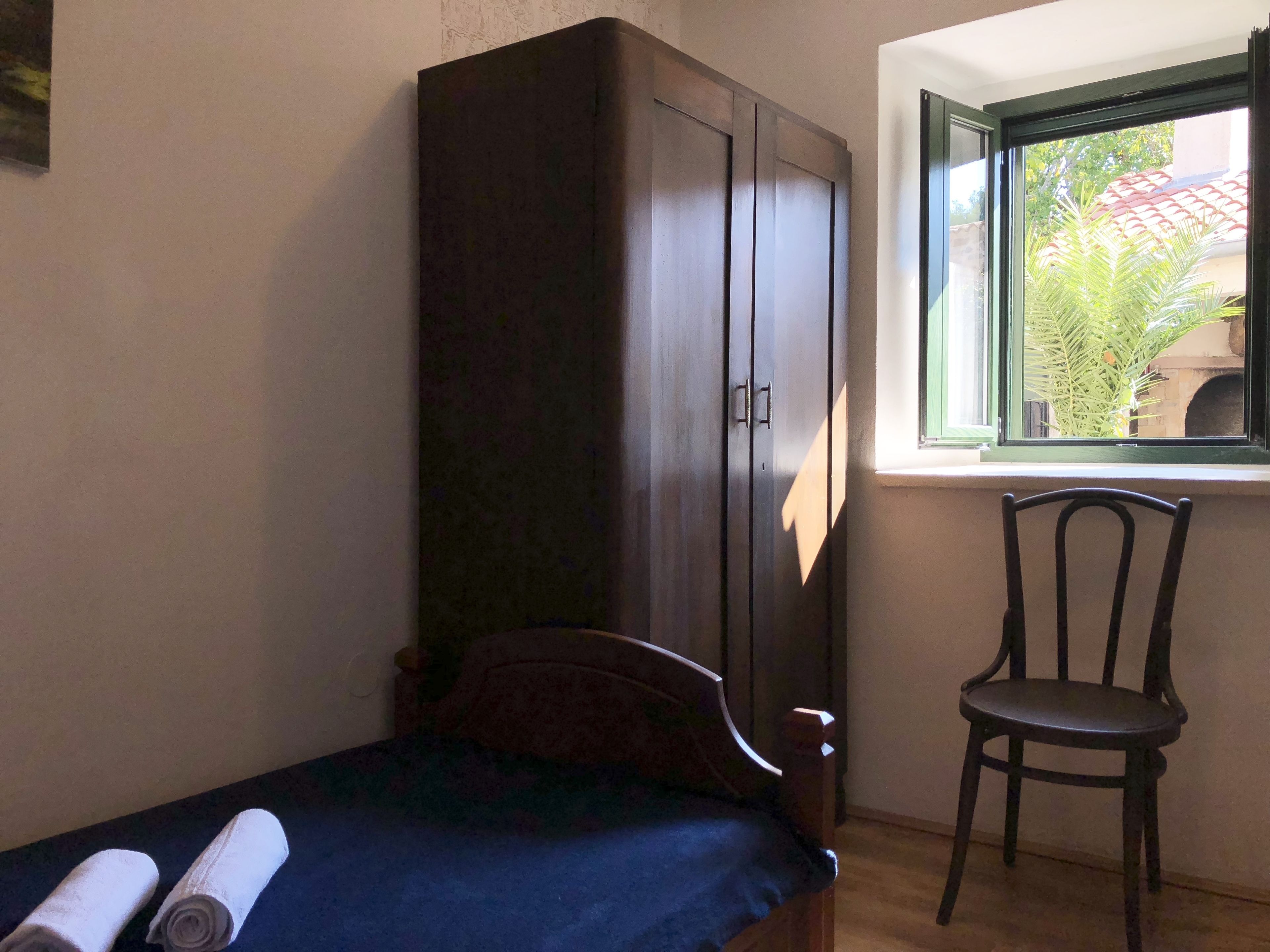 Una habitación pequeña con cama, armario de madera, una silla y ventana con vistas a espacios verdes.
