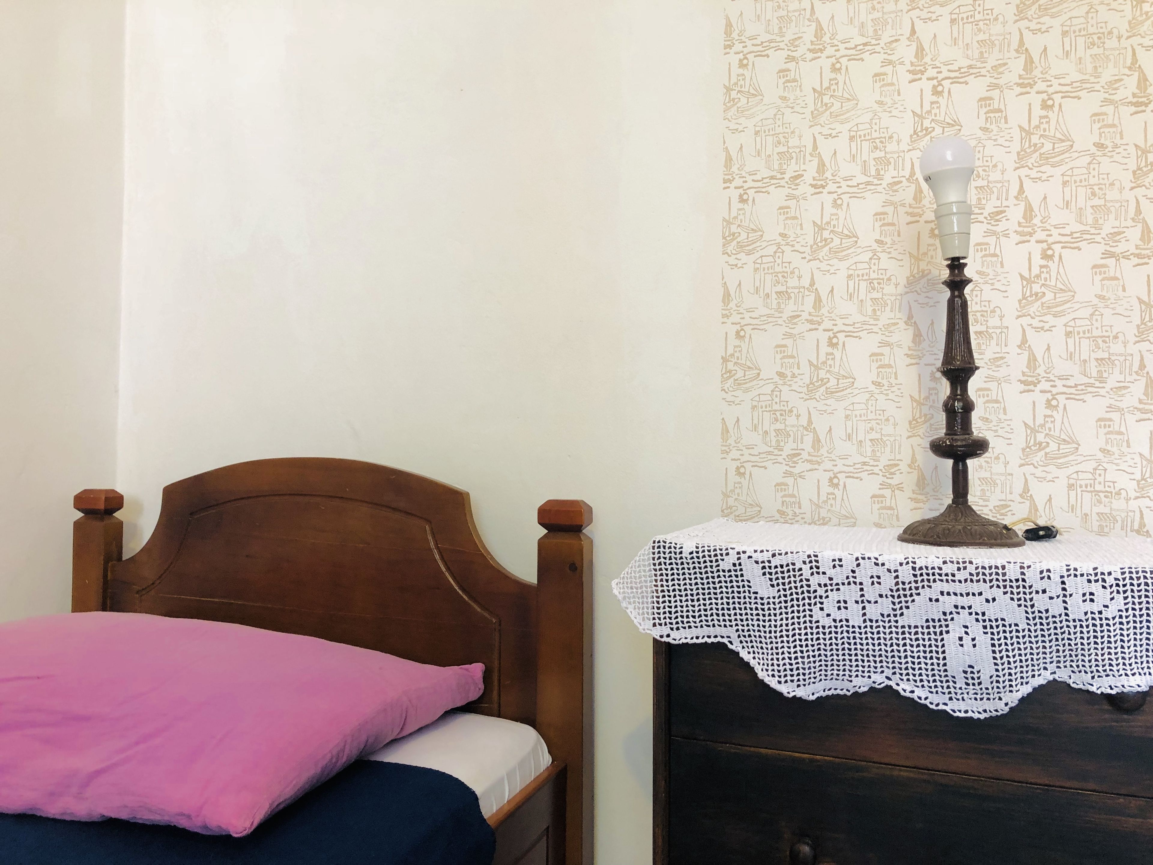 A bedroom with a wooden bed, a pink pillow, a white blanket, a lace tablecloth, and a wall with a nautical pattern.